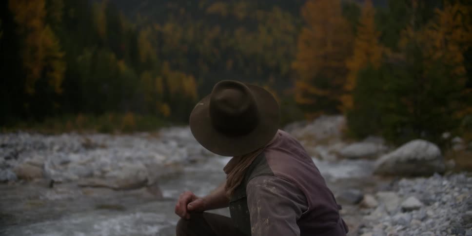Male prospector throws gold panning plate into water then stands to look toward mountains towering over nearby conifer forest