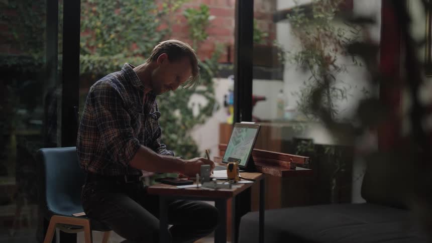 A man is sitting at a desk looking at a laptop computer and writing on paper as his phone sits on the desk
