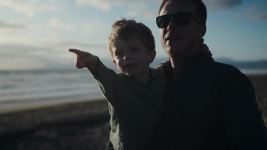 A man carries a boy in his arms as the two of them point at their surroundings on a beach during the day
