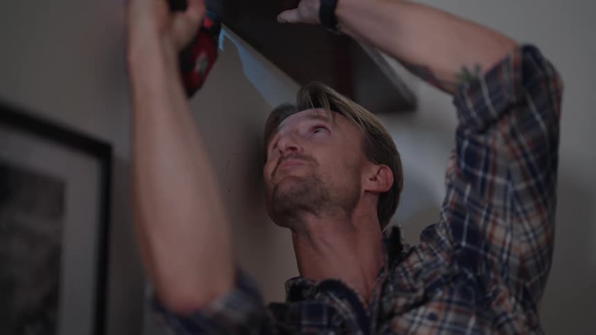 A blond bearded man wearing a long sleeve plaid shirt and watch drills a wooden shelf above him