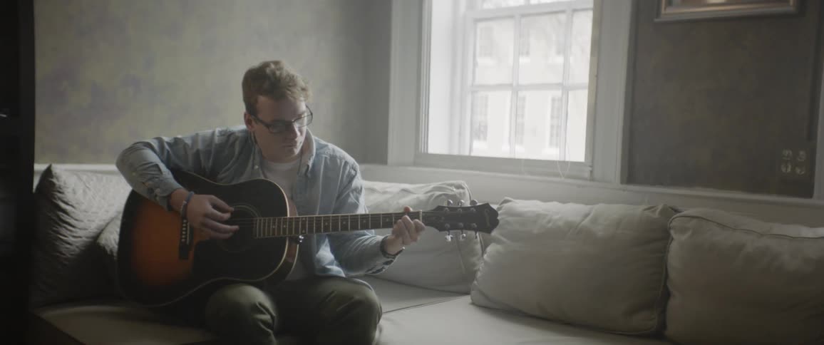 A bespectacled young man plays an acoustic guitar near a georgian window