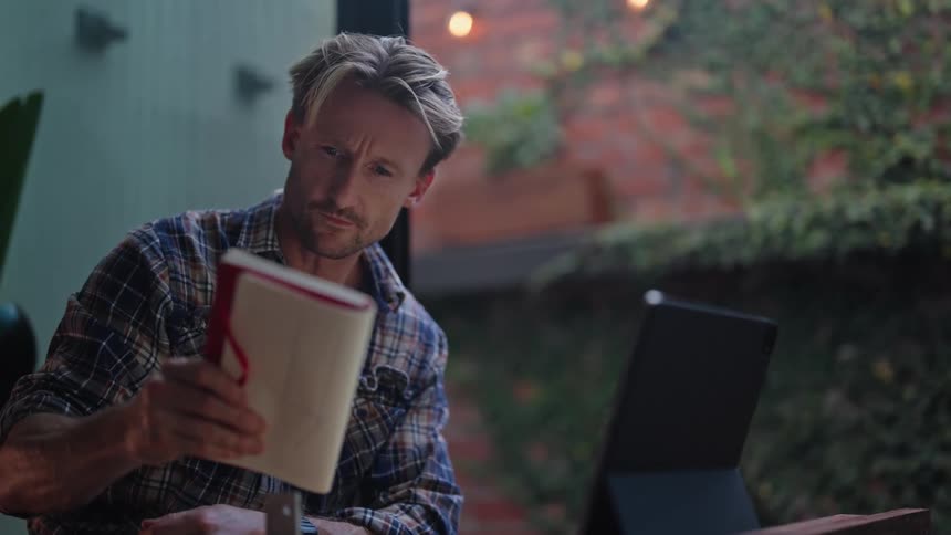 Man sitting at a table looking at a book then turning to his tablet to write on it