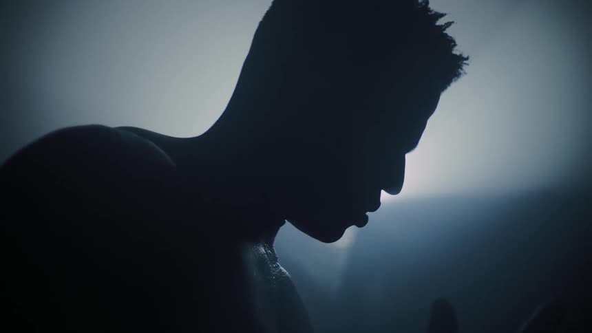A shirtless man with a tense expression making dramatic hand movements while looking down with flashes of light illuminating his face
