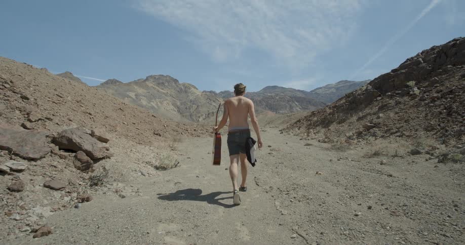 A man holding a guitar walks towards some hills ahead