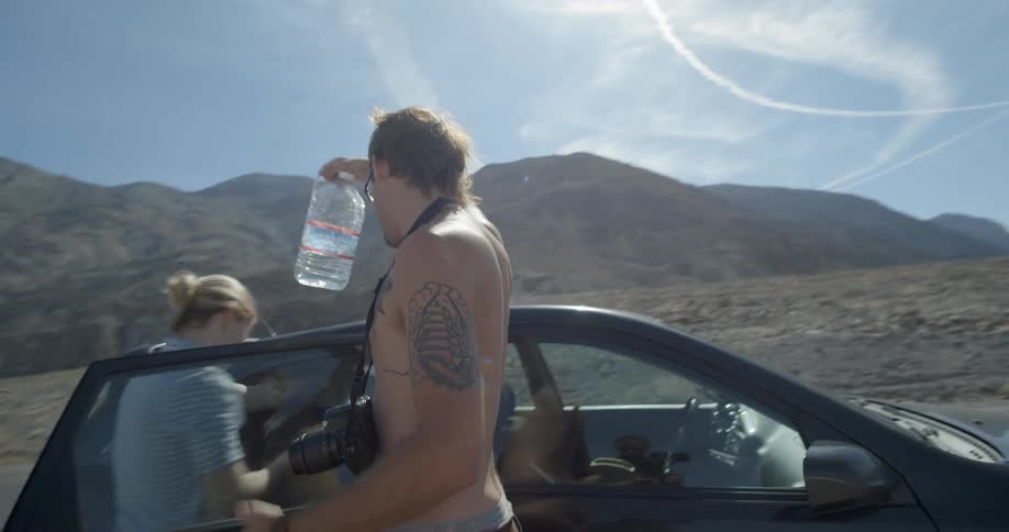 Man drinks out of a large water bottle outside a car in the desert
