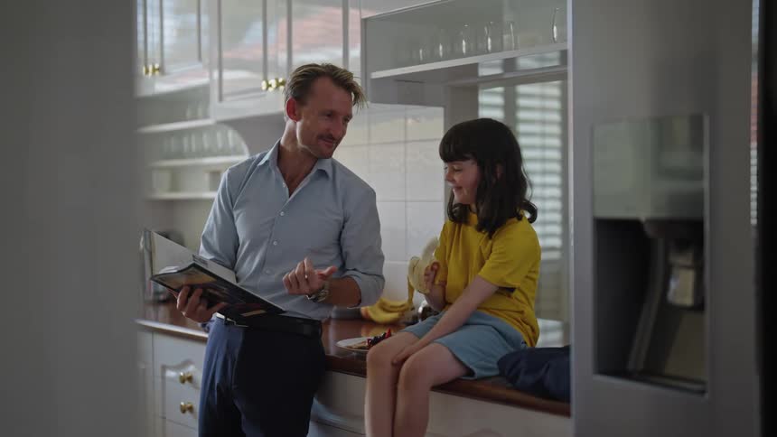 Daughter eats a banana while father shows her a book in the kitchen