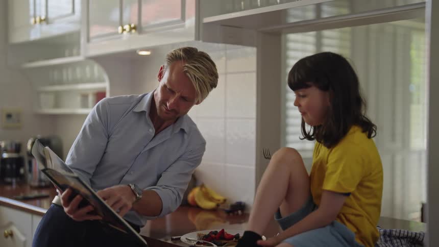 Daughter sits on kitchen counter eating while she and her father discuss book contents
