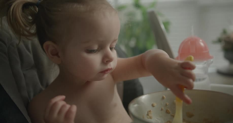 Toddler sitting in a high chair eating food with a spoon from a bowl