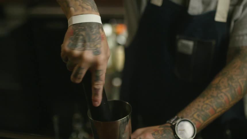 A bartender uses a muddler to mash something in a tin