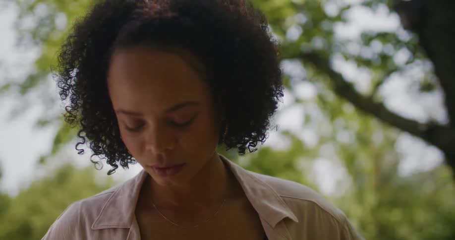 A close up of a woman standing outside under a tree while looking down around her feet