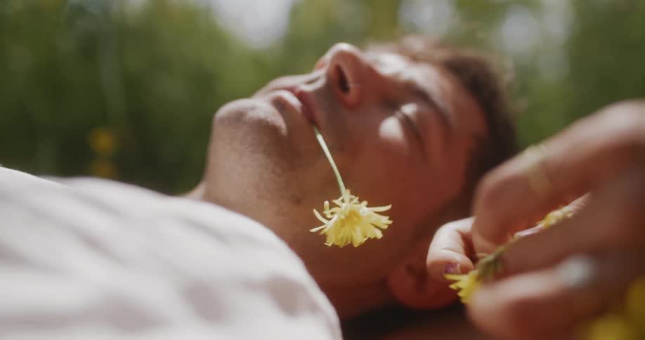 Woman plays with flowers next to man lying on his back on ground with flower in his mouth