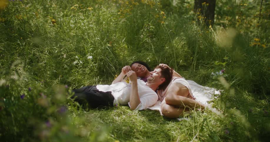 Man and woman laying together on a blanket in a grass field under a tree on a sunny day