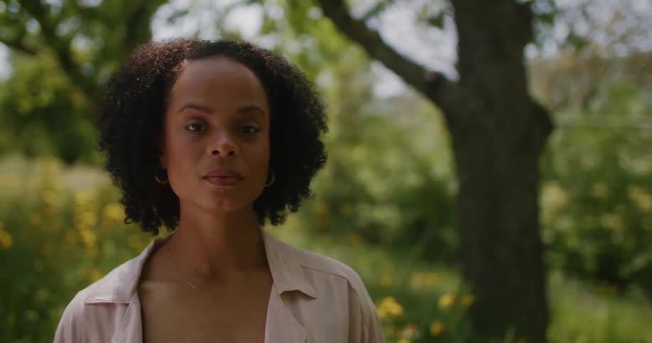 A woman standing beneath a tree in a field raises her head to look into the camera for a moment before grinning