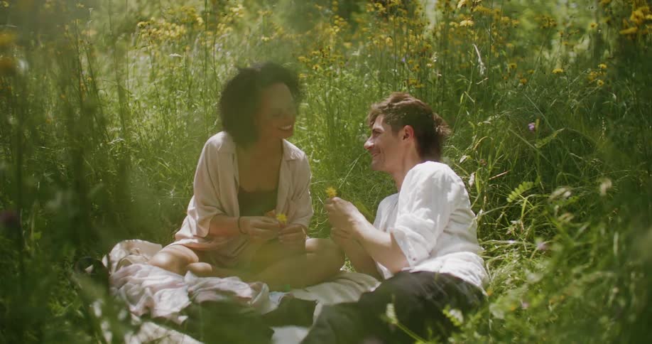 A man and woman sitting outside in the grass of a field laughing and smiling holding flowers in their hands