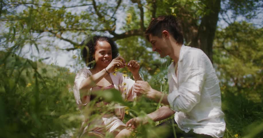 A man and woman sitting in a field outdoors tying flowers together and laughing and smiling