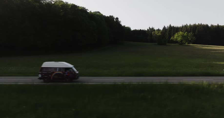 Van drives along highway through field bordering conifer woods