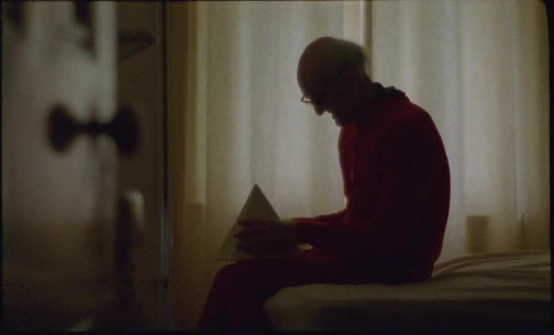 Elderly man sitting on a bed with a ceramic pyramid on his lap