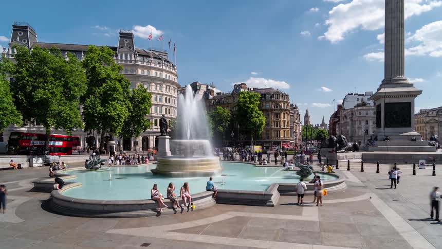 Crowd of pedestrians moves around large fountain in plaza near historic buildings on cloudy day