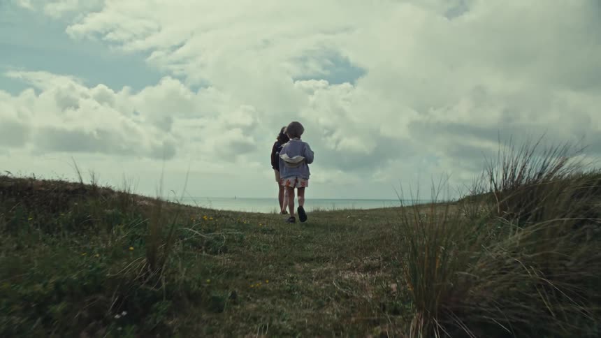 A little child running toward their mother next to the beach and stops to look over the edge of the coast