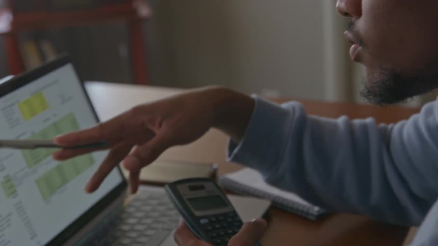 A man points at the laptop and uses his calculator