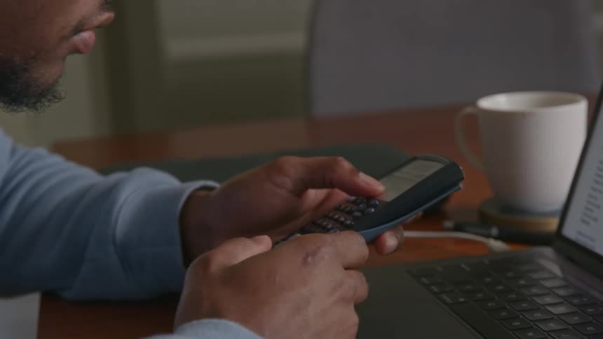 Close up of a man using a calculator while sitting in front of a laptop on a table