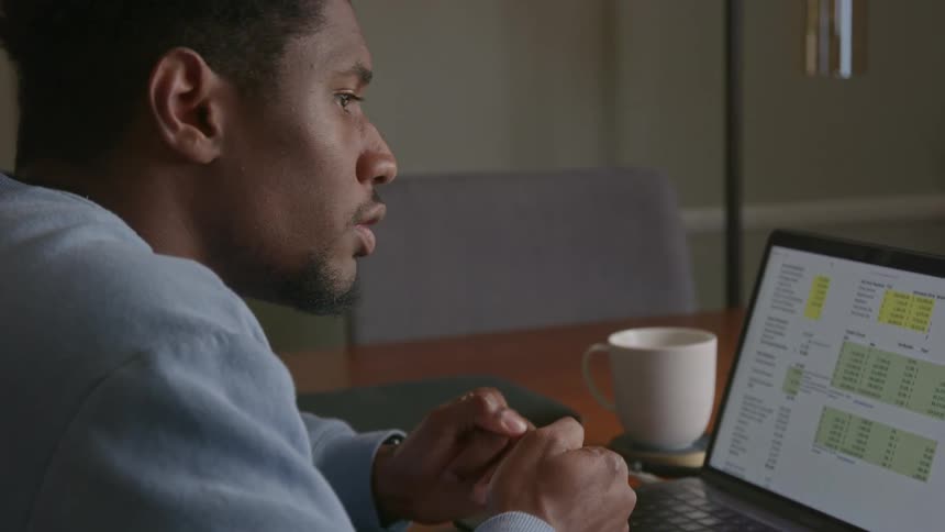 Close up of a man sitting in front of a laptop while typing into a calculator in his hands before making a frustrated face