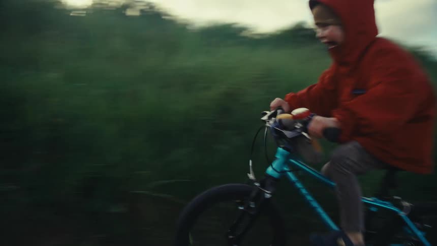 Young child riding a bicycle on a dirt road while someone walks behind him
