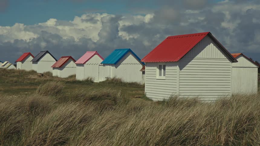 Series of small houses with colored roofs which include red and blue no fence and surrounded with grasses on a windy day