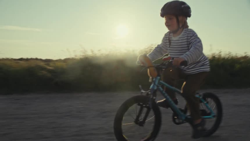 A young boy riding his bicycle past his mother who is next next to him as the sun sets
