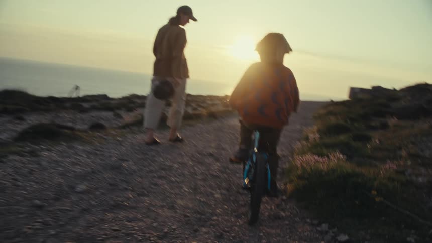 A child riding their bike behind their mother walking in front of them on a trail near the beach as the sun sets