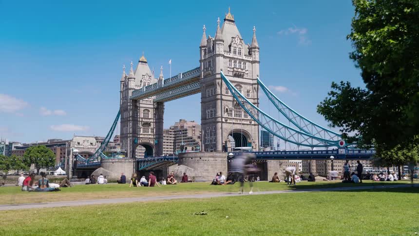 A crowd sits in a park below a city bridge