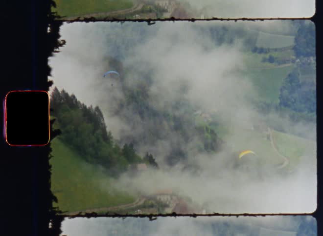 An aerial of two people sailing through the sky on parasails over cloudy grounds with houses built into the hillsides