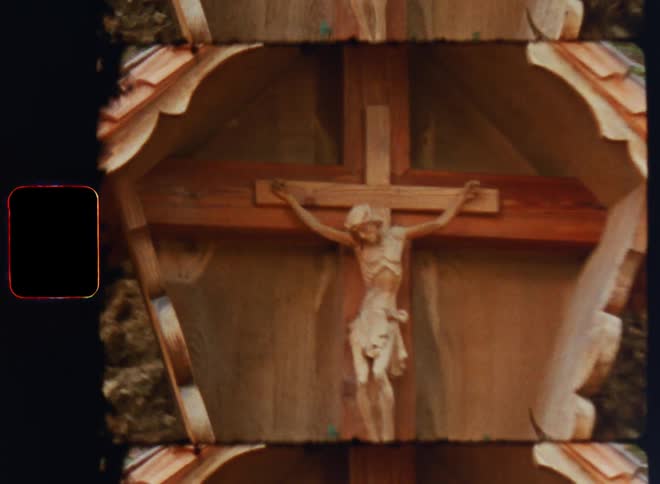 A wood carving of jesus christ on the cross is house in a wooden frame with a plant growing below placed in a forest