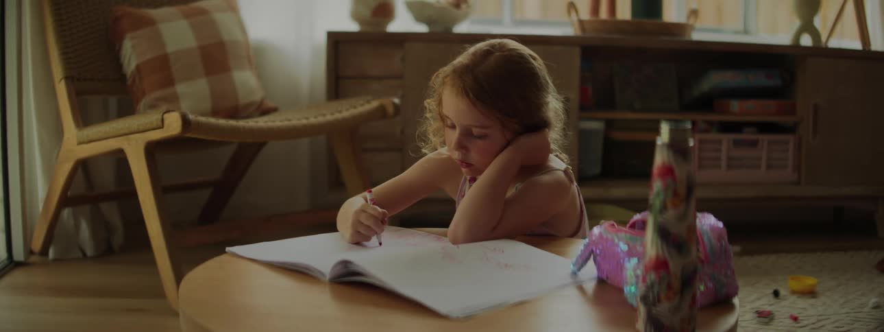 A little girl in a room seated on the floor in the room and writing with a pencil on the book on the table