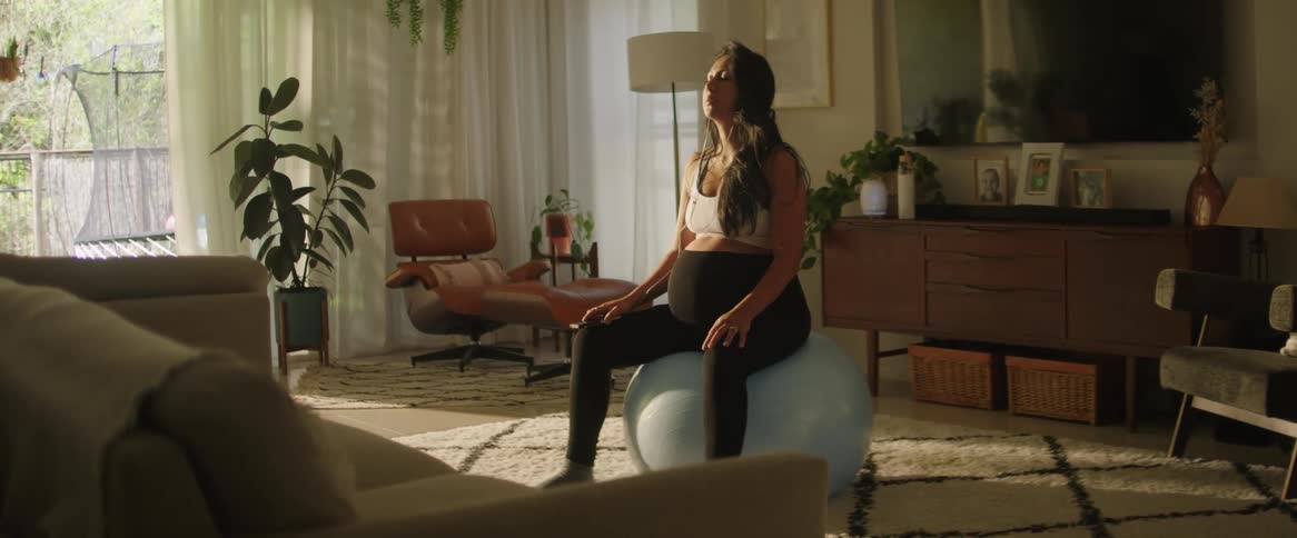 A pregnant woman meditates atop a yoga ball in the living room of a house during the day