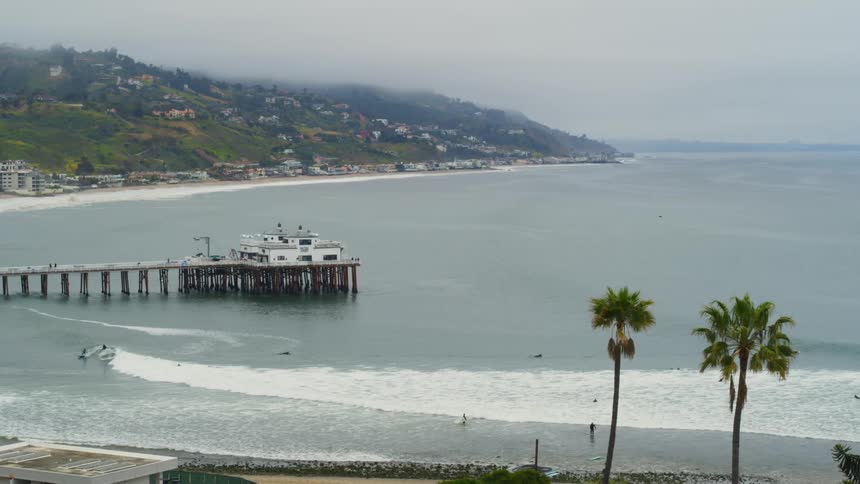 People are surfing in the water near a building at the end of a pier with buildings on the hillside along the coast