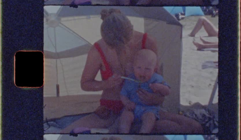 Adult woman at the beach feeding her smiling baby food while other people lying down sunbathe on the background