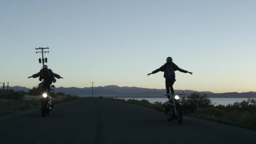 Two people stand on their motorcycles while riding them