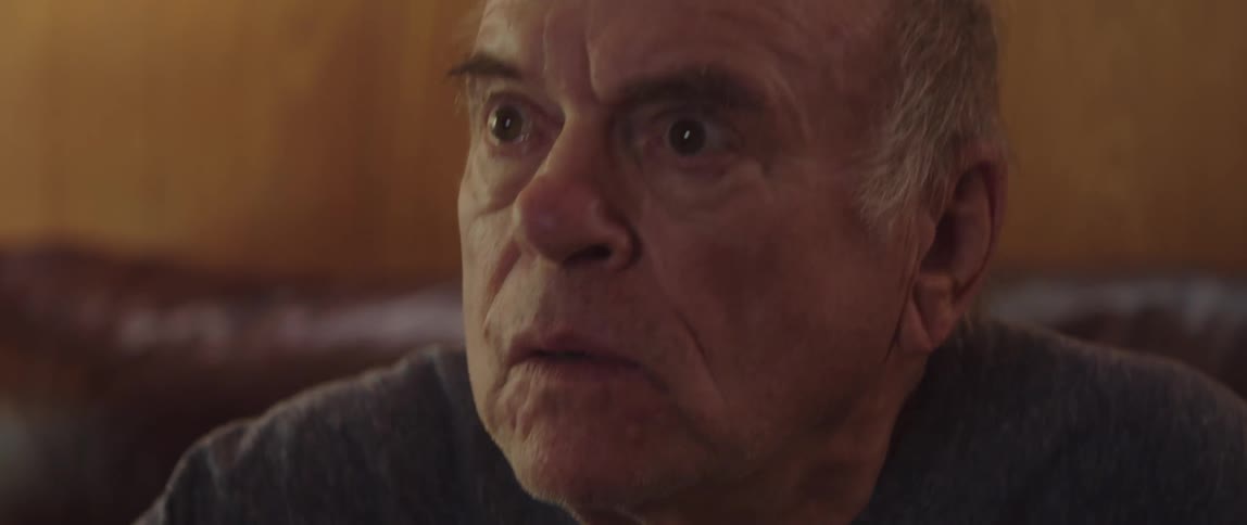 A man is smiling and sitting on his couch while watching tv