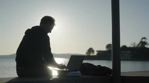 A man is typing on his keyboard by the water