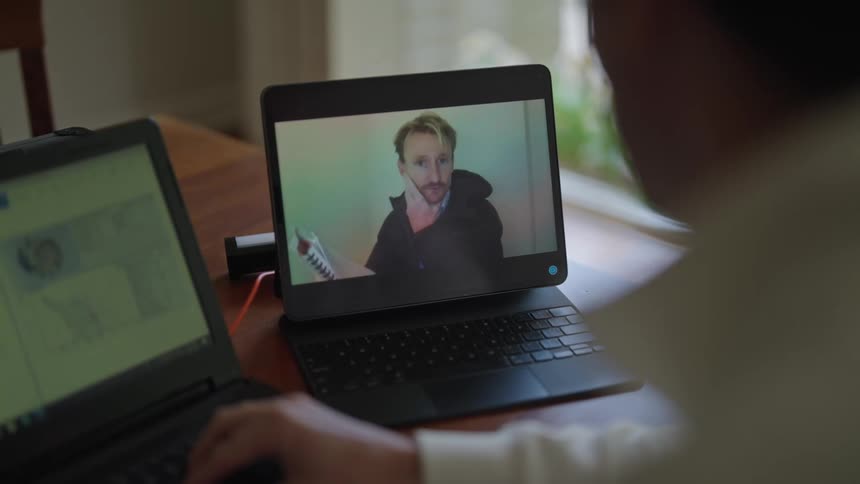 Man working on his desk having a video call with another man on one of his laptops while typing on the other one