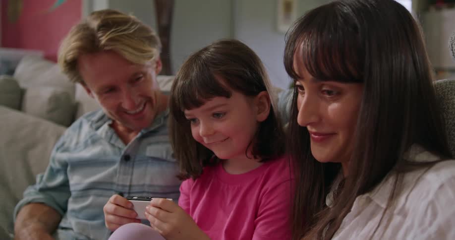 A family of a mother father and daughter are sitting on the couch while the daughter plays a handheld video game