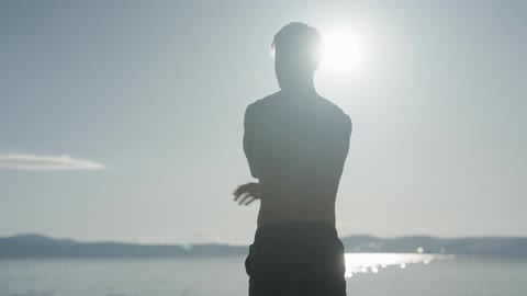 Shirtless man standing in front of the sea stretching his arms while admiring the landscape on a sunny day