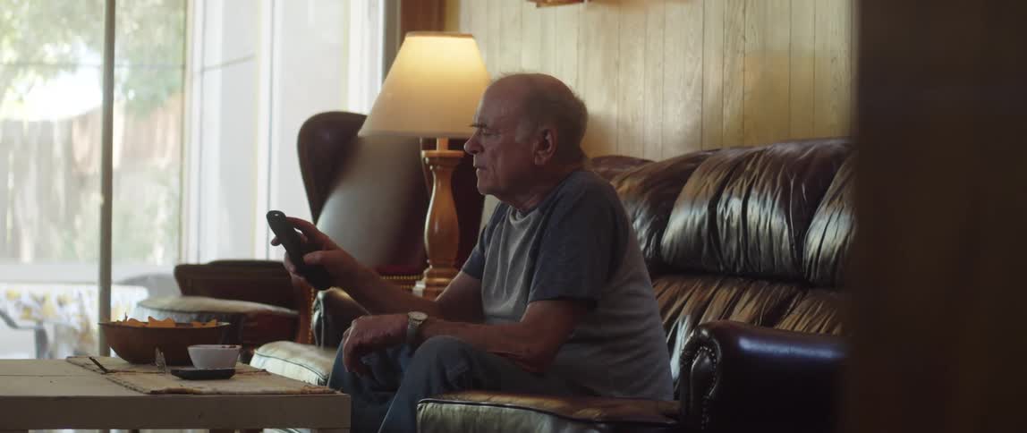An old man sitting on the living room couch watching tv reaches for the remote