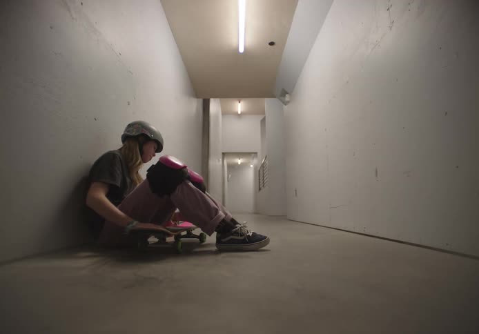 A skateboarder fidgets while sitting against a wall in a corridor