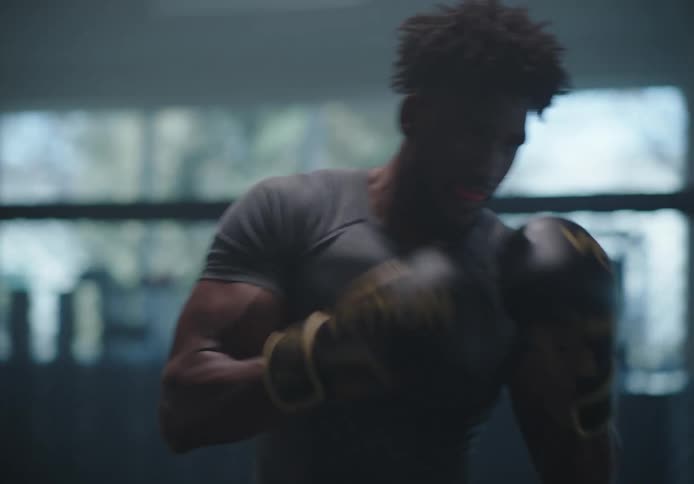 A muscular man wearing boxing gloves spars with another person in a gym