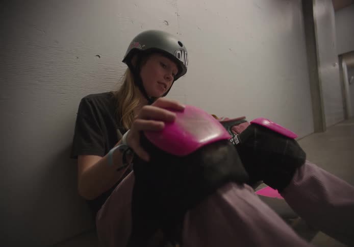 A female skateboarder removes her knee pads while sitting against a wall in a corridor