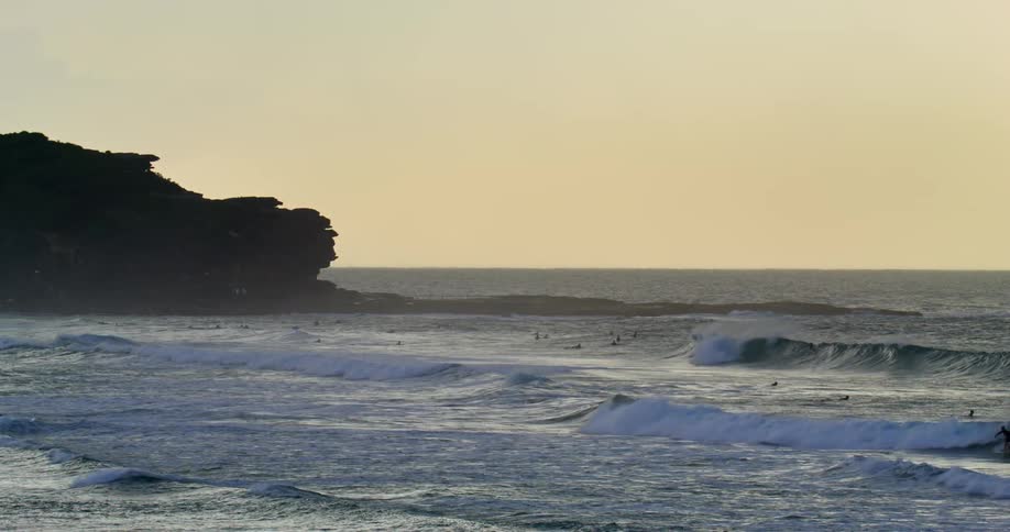 A large group of people are swimming or surfing in the ocean at sunset