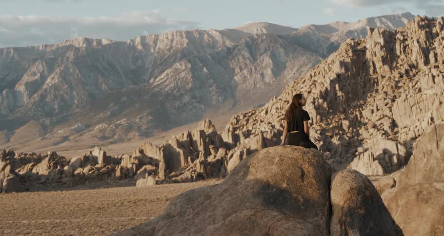 A woman sits atop a rock formation in a mountainous area of a desert during the day
