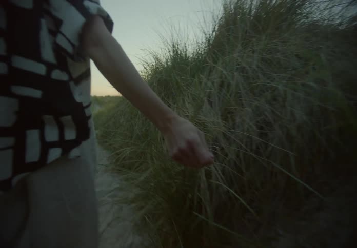 A woman reaches out and touches the tall grass as she walks towards the shoreline